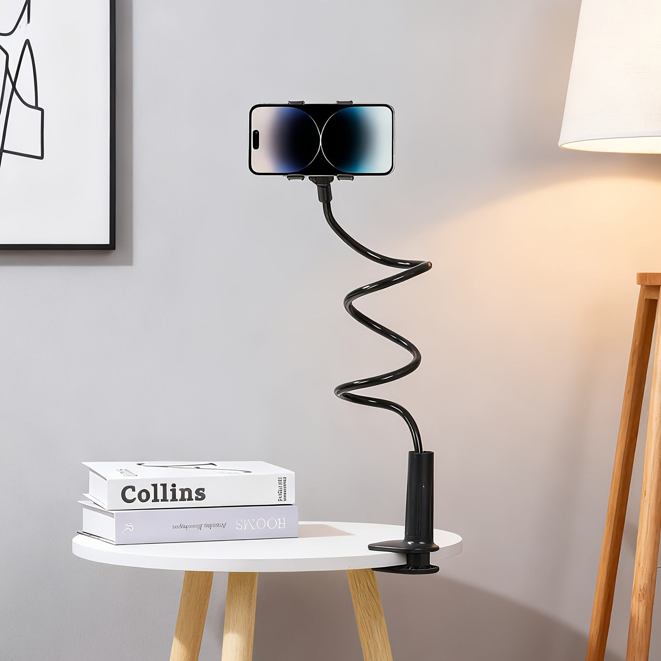 Black ABS flexible gooseneck phone holder with a clamp base on a white side table, holding a smartphone. 360° adjustable.